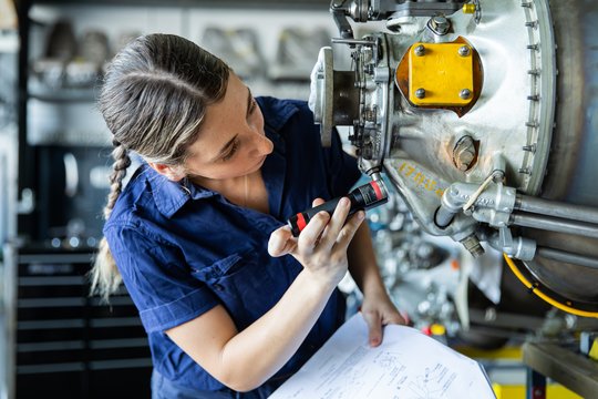 Eine junge Frau an ihrem Arbeitsplatz im Flugzeugbau. Sie hält ein Werkzeug in der Hand und arbeitet an einem Metallteil. 