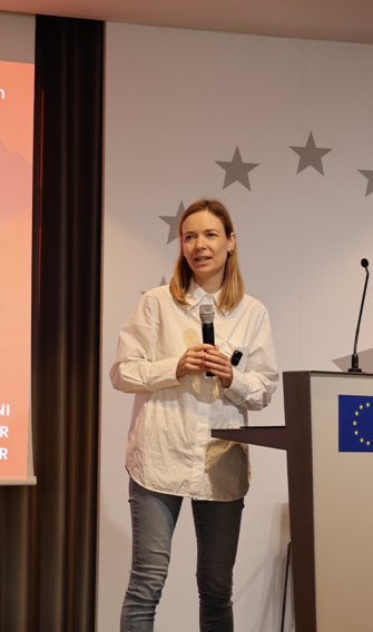Eine blonde Frau am Redepult, im Hintergrund einige Sterne der europäischen Flagge.