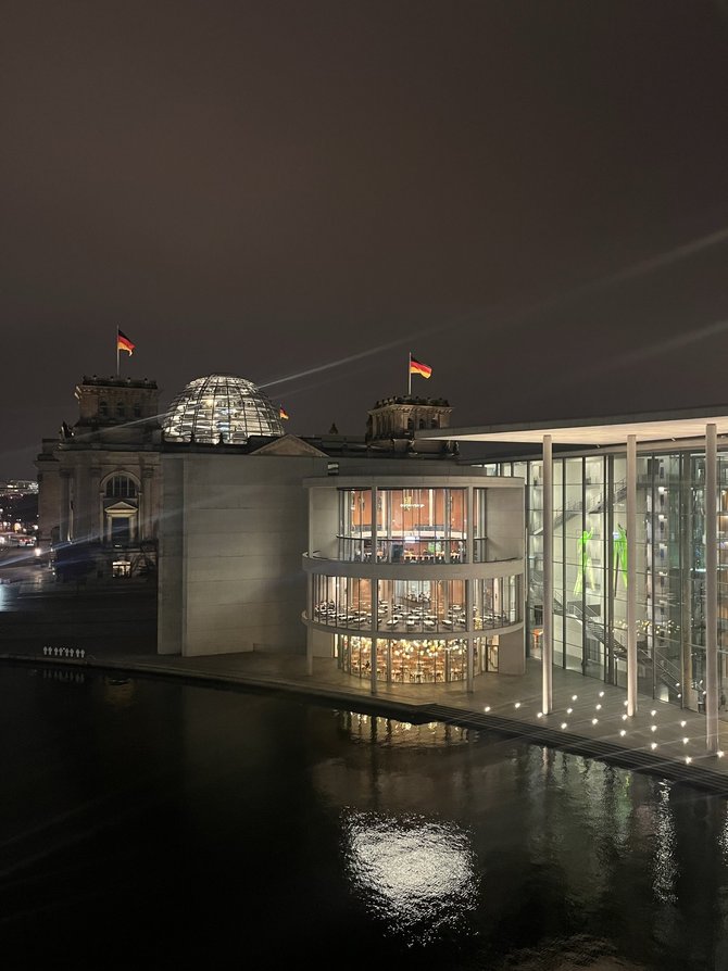 Nachtaufnahme des Berliner Regierungsviertels mit Blick über die Spree auf ein modernes, verglastes Parlamentsgebäude mit mehreren runden Etagen; im Hintergrund ist das historische Reichstagsgebäude mit beleuchteter Glaskuppel und mehreren wehenden Deutschlandfahnen zu sehen, das Wasser im Vordergrund spiegelt die Lichter.