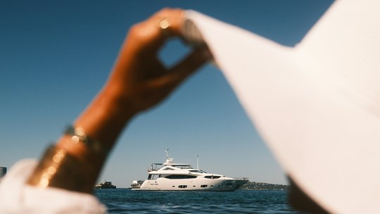 Eine weiße Motoryacht liegt auf dem Meer, im unscharfen Vordergrund formt eine Hand mit Goldarmbändern einen Kreis aus Daumen und Zeigefinger.