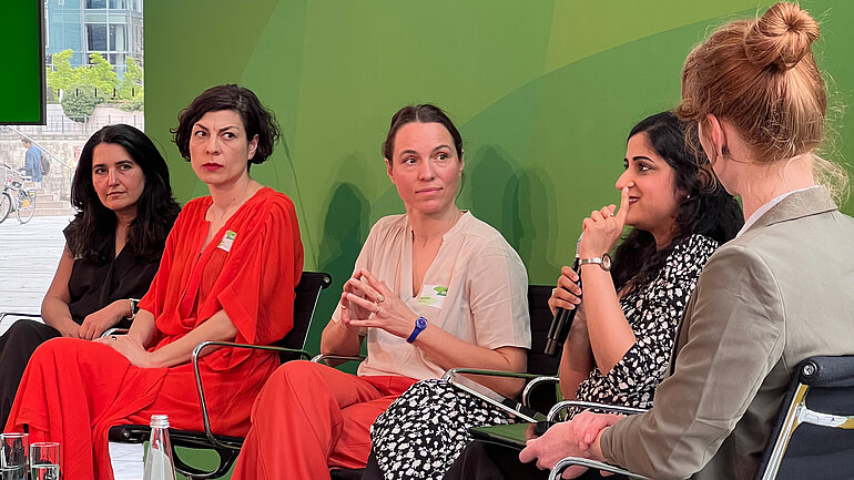 Fünf Frauen sitzen auf Stühlen nebeneinander vor einer grünen Wand