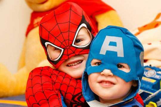 Zwei Kinder verkleiden sich als Superhelden und Superheldinnen. Eines trägt eine rote Verkleidung, das andere eine blaue Maske, sie lachen in die Kamera. 