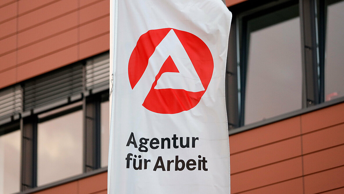 Weiße Flagge mit dem Logo der Agentur für Arbeit vor einer Hausfassade.