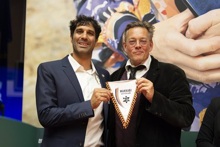 zwei Männer mit Wimpel in der Hand, auf dem Makkabi steht, vor einem Ausschnitt des Bühnenhintergrunds