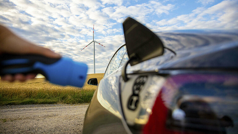 Eine Hand hält einen Ladestecker an die Steckdose eines Elektroautos. Im Hintergrund ein Getreidefeld, ein Windrad und blauer Himmel. 