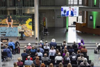 Im Vordergrund sitzen zahlreiche Teilnehmende, sie schauen auf einen Videobildschirm, wo Dominik Krause zu sehen ist.