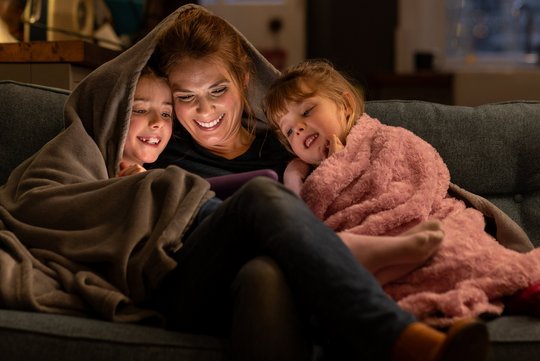 Eine Frau sitzt mit ihren zwei Kindern eingekuschelt unter einer Decke auf dem Sofa