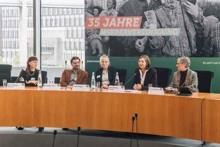 Die 5 Panel-Gäste sind zu sehen, hinter ihnen ist auf einem 3 Meter hohen Aufsteller das Motiv der Veranstaltung zu sehen: Eine schwarz-weiß Aufnahme kurz nach dem Mauerfall, die im Vordergrund zwei sich küssende Menschen mit je einem Geldschein zeigt 