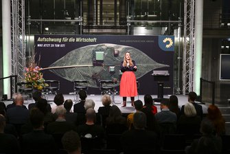 Eine Frau mit wallendem Rock und blonden Haaren spricht auf der Bühne