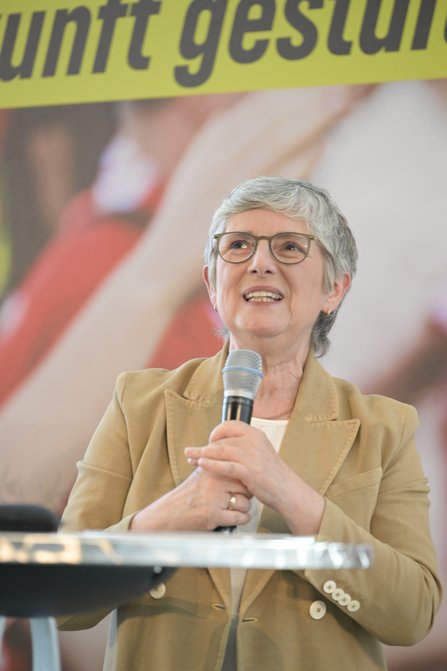 Frau mi Mikro in der Hand, im Hintergrund ein verschwommenes Bühnenbild, vorne im Anschnitt ein silberner Stehtisch 