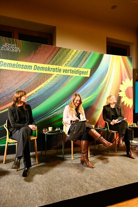 Drei Frauen sitzen auf einer Bühne, im Hintergrund steht ein Banner mit der Aufschrift "Gemeinsam Demokratie verteidigen"