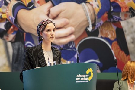 Helen Braun am Redepult, im Hindergrund das Motiv des Abends: Eine Frau, die ein Gebetbuch in der Hand hält und ein buntes Kleid trägt. Nur der Oberkörper ohne Kopf ist sichtbar.