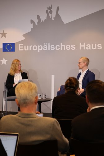 2 Personen auf der Bühne sind in einer Interviewsituation. Im Vordergrund Rücken und Köpfe von Teilnehmenden.