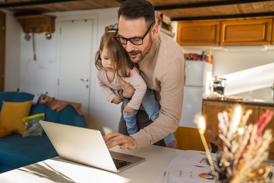 Ein Vater steht zu Hause in der Küche, hat sein Kind auf dem Arm und tippt etwas in den Laptop