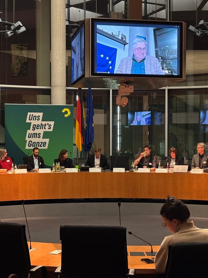 Großer Konferenzsaal mit halbrunder Tischanordnung; vorne sitzen mehrere Personen an Mikrofonen, links ein grünes „Uns geht’s ums Ganze“-Plakat, über ihnen hängt ein großer Bildschirm, auf dem Martin Häusling in Videokonferenz vor einer EU‑Flagge zu sehen ist, im Vordergrund sitzt mit dem Rücken zur Kamera eine Person an einem Einzelplatz und macht sich Notizen.