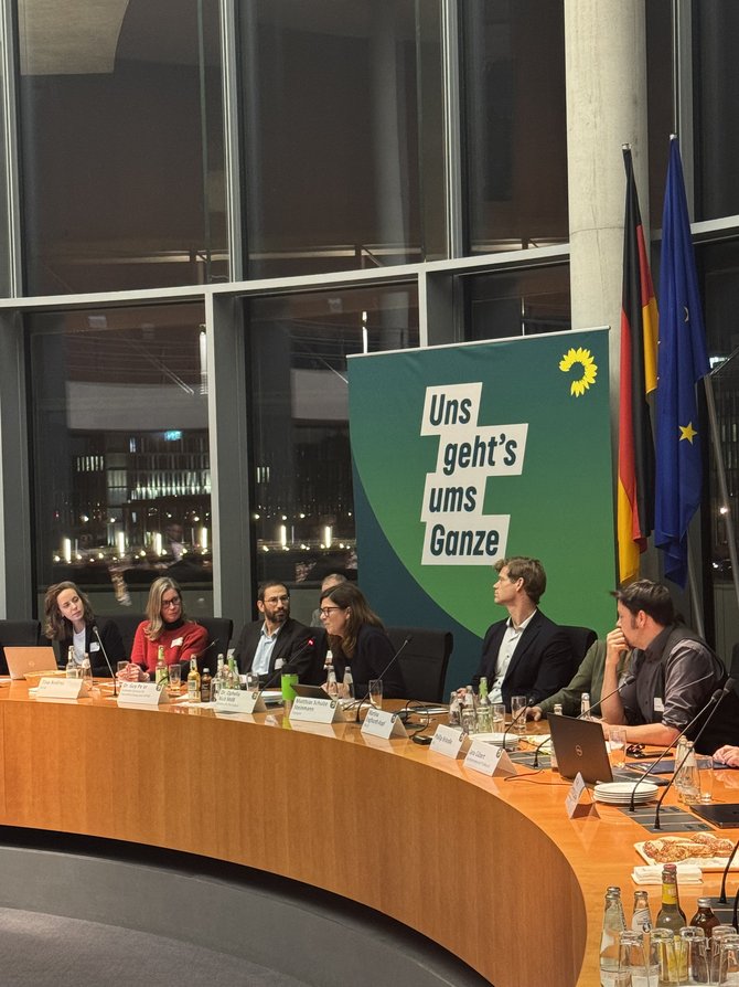 Runder Sitzungstisch in einem Glas-Saal, an dem etwa zehn Personen sitzen; hinter der zentralen Sprecherin steht ein großes grünes Banner der Bundestagsfraktion Bündnis 90/Die Grünen mit der Aufschrift „Uns geht’s ums Ganze“, daneben Deutschland- und EU‑Flagge, auf dem Tisch liegen Mikrofone, Getränke und Unterlagen.