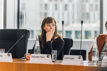 Prof. Hedwig Richter sitzt am Tisch im Podium, Namensschilder, Mikrofone und Wasserflaschen- und Gläser sind zu sehen
