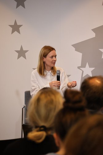 Eine blonde Frau sitzt vor dem grauen Bühnenhintergrund und spricht ins Handmikrofon.