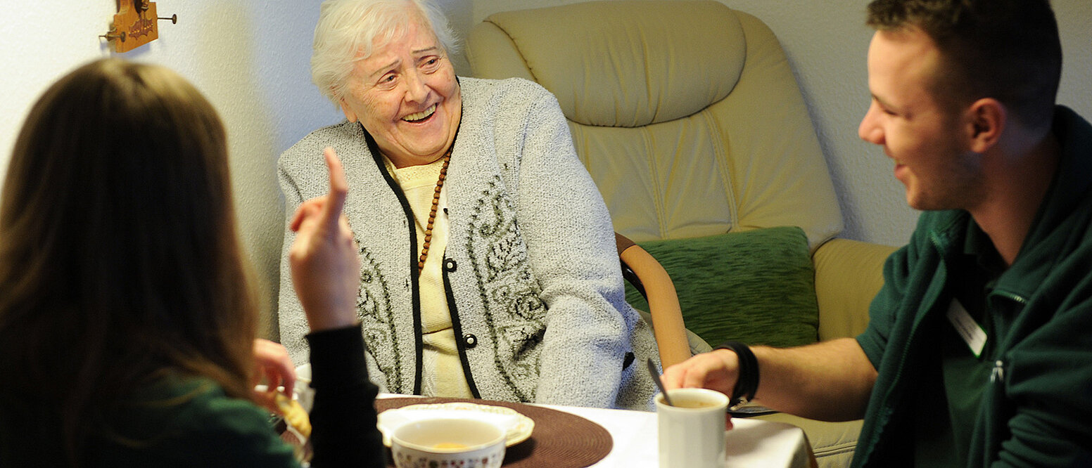 Zwei  Bundesfreiwilligendienstleistende besuchen im Rahmen ihres Freiwilligendienstes beim Pflegedienst pro vita eine Seniorin und leisten ihr Gesellschaft. Gemeinsam sitzen sie am Tisch und trinken Kaffee. 