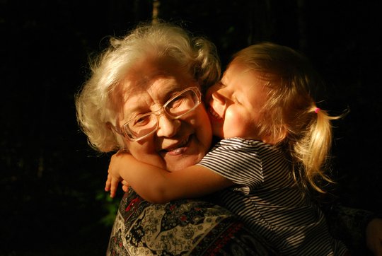 Ein lachendes Kleinkind mit blondem Zopf umarmt eine ältere Frau mit weißem Haar und Brille, die in die Kamera lächelt.