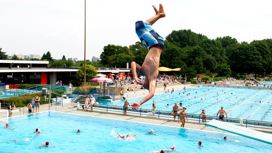 Blick in ein Freibad. Im Vordergrund sind man einen jungen Mann mit einem Salto ins Wasser springen