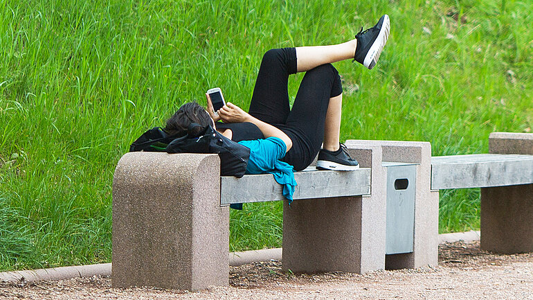 Eine Frau liegt auf einer Parkbank und guckt in ihr Smartphone.