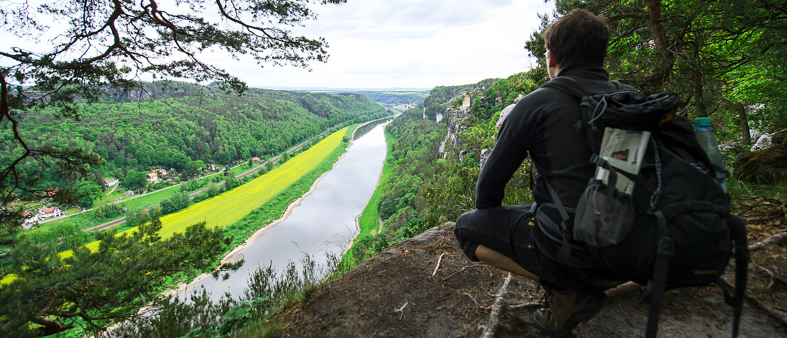 Ein Wanderer sitzt auf einem Felsen in der Nationalparkregion Sächsische Schweiz und schaut ins Elbtal hinab.