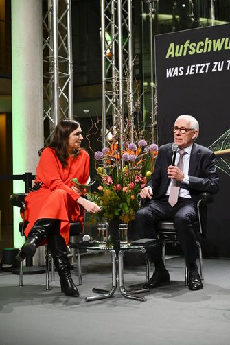 Moderatorin und ein Gast unterhalten sich auf der Bühne, zwischen ihnen ein großes Blumengestecl