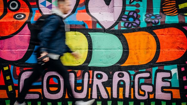 Courage Ein Mensch geht eine Grafittiwand mit dem Schriftzug "Courage" entlang