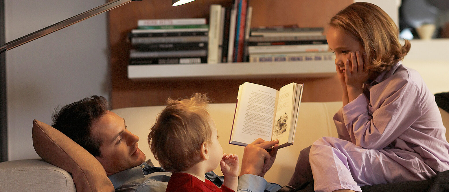 Eine Vater liegt auf einem Sofa und liest seinen zwei Kindern aus einem Buch vor