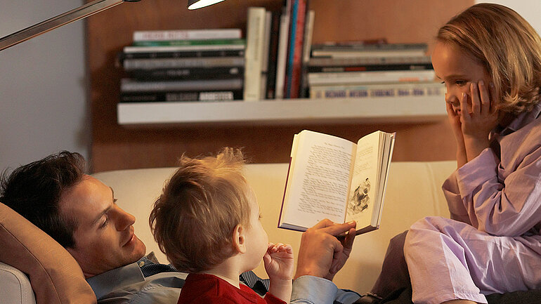 Eine Vater liegt auf einem Sofa und liest seinen zwei Kindern aus einem Buch vor