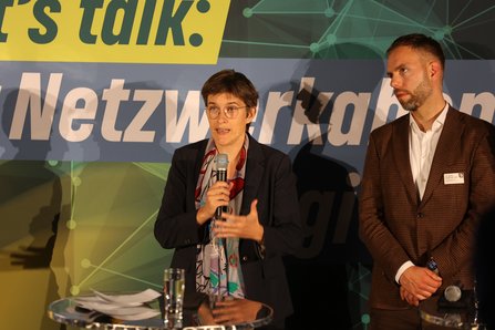 Frau mit Mirko vor Bühnenhintergrund, rechts ein Mann, der zu ihr blickt