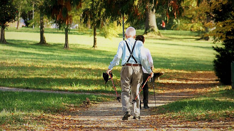 Zwei ältere Menschen von hinten, gehen auf einem Sandweg durch einen Park. 