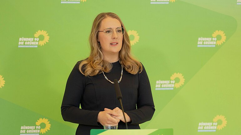 Katharina Dröge vor einer grünen Rückwand mit dem Logo der grünen Bundestagsfraktion.