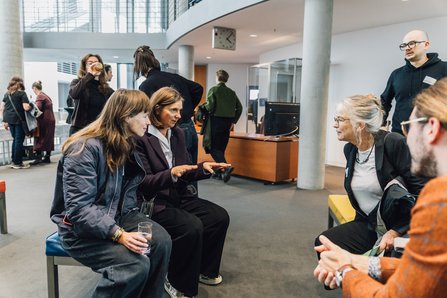 4 Personen sitzen auf Polsterbänken im Foyer des Europasaals und unterhalten sich, daher stehehend weitere Personen in Gesprächen