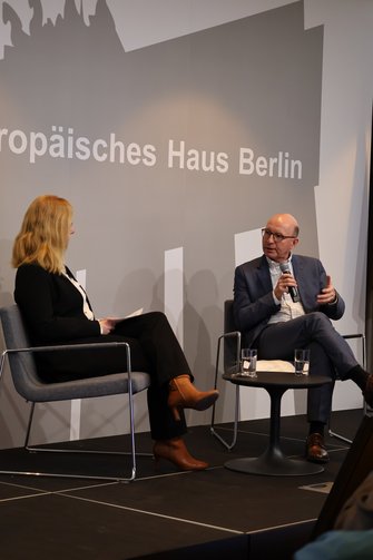 Eine Frau und ein Mann in einer Interviewsituation auf der Bühne. Auf dem Bühnenhintergrund ist das Brandenburger Tor zu erkennen, davor steht "Europäisches Haus Berlin".