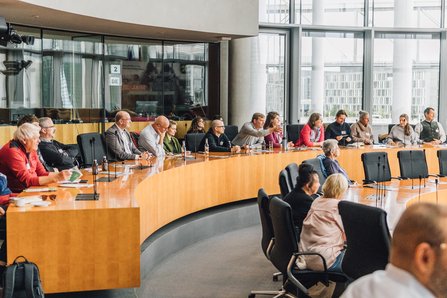 Das Bild zeigt Veranstaltungsgäste, die im Europasaal an einem großen runden, parlamentarischen Tisch sitzen