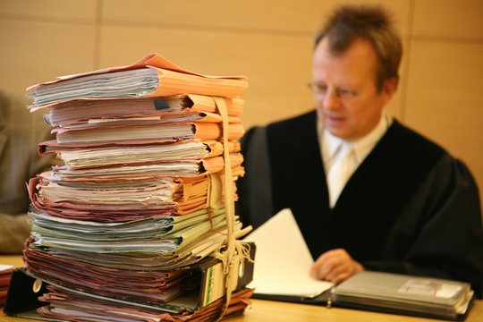 Gerichtsakten stapeln sich auf einem Tisch in einem Gericht, dahinter sitzt ein Richter und blättert in einer Akte.