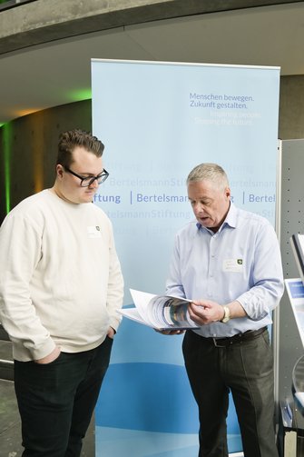 Zwei Männer schauen gemeinsam in eine Publikation, im Hintergrund ein RollUp der Bertelsmann-Stiftung.