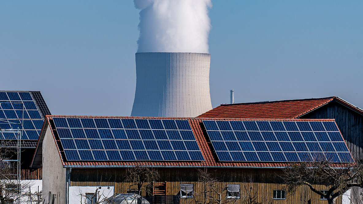 Wasserdampf steigt aus dem Kühlturm vom Atomkraftwerk Isar 2, davor steht ein Stall mit einer Photovoltaikanlage.
