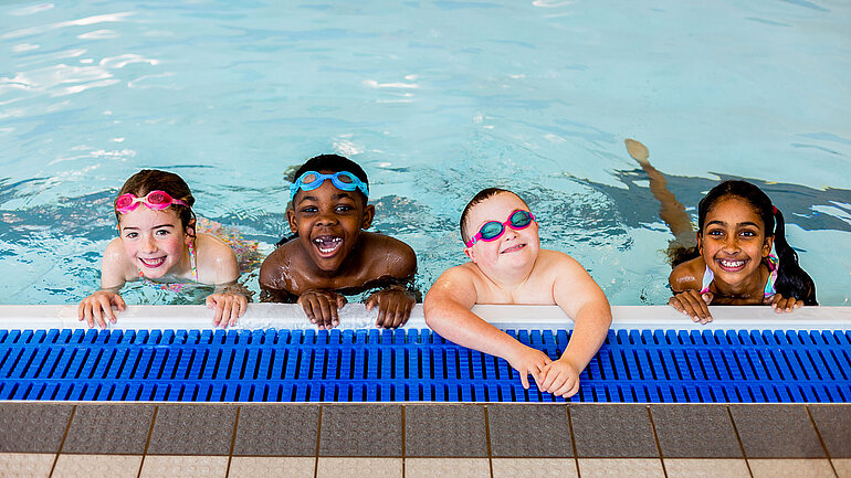 Vier Kinder gucken über den Rand eines Schwimmbads.
