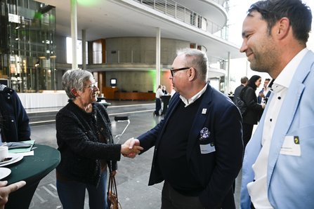 Frau begrüßt mit Handschlag einen Mann. Im Vordergrund rechts ein weiterer Mann.