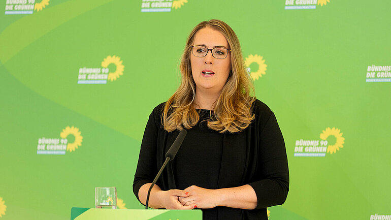 Katharina Dröge steht am Mikrofon vor einer grünen Wand mit vielen Logos der grünen Bundestagsfraktion.