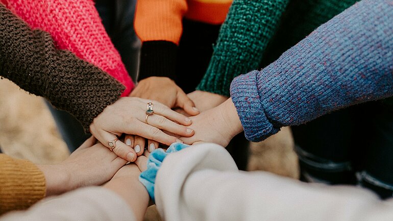 Viele Personen stehen im Kreis und legen ihre Hände übereinander. Sie haben bunte Pullover an.