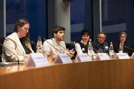 Junger Man (Ron Dekel) guckt intensiv in das Publikum (nicht sichtbar), neben sich andere Podiumsgäste