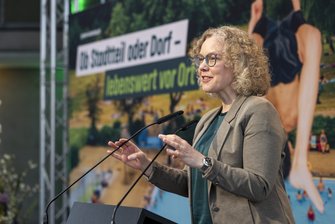 Eine Frau am Redepult, auf dem Bühnenhintergrund steht der Slogan des Konferenz "Ob Stadtteil oder Dorf - lebenswert vor Ort"
