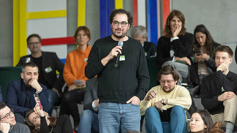 Regisseur Can Tanyol spricht, im Hintergrund sitzt das Publikum.