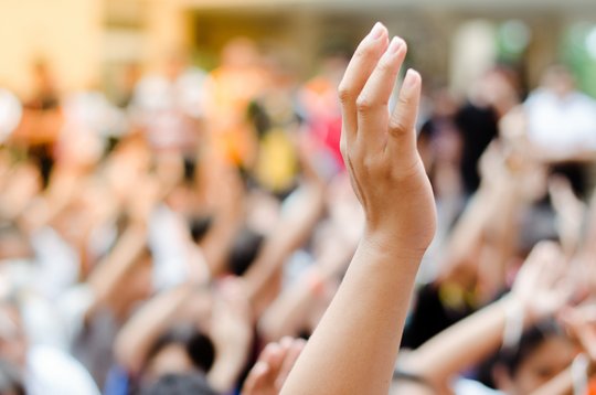 Ein unscharfes Bild in eine Menschenmenge, viele Menschen heben ihre Hände. Eine Hand ist scharf im Vordergrund zu sehen.