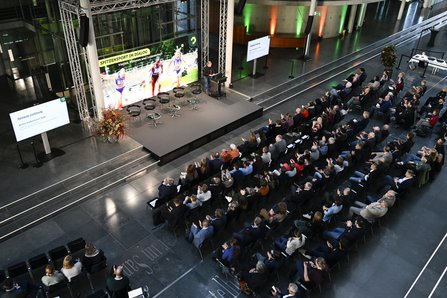 Blick von oben in die Veranstaltungshalle, Menschen in Stuhlreihen vor Bühne, dort ein Frau am Redepult