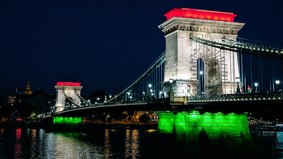 Ein Fluß mit einer großen Brücke, die in der Nacht rot, weiß und grün angeleuchtet wird.